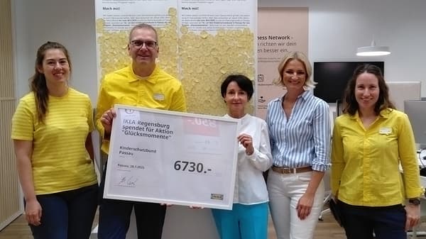 5 Personen stehen vor einer Wand mit aufgeklebten Blüten. 2 Personen halten einen Spendenscheck in den Händen mit einem Spendenbetrag von 6730€, den IKEA Regensburg an den Kinderschutzbund in Passau spendet.
