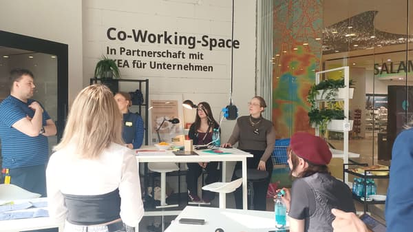 7 Personen stehen im IKEA Co-Working-Space an Tischen im Kreis- Zwei davon tragen IKEA Kleidung. Es liegen verschiedene Arbeitsmaterialien auf den Tischen.