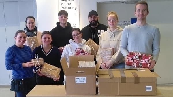8 Personen stehen vor IKEA Kartons und Paketen und halten eingepackte Geschenke in den Händen. 