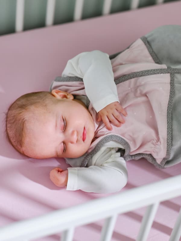 A baby in a pink sleep bag on a pink fitted sheet is asleep in a white GULLIVER crib.