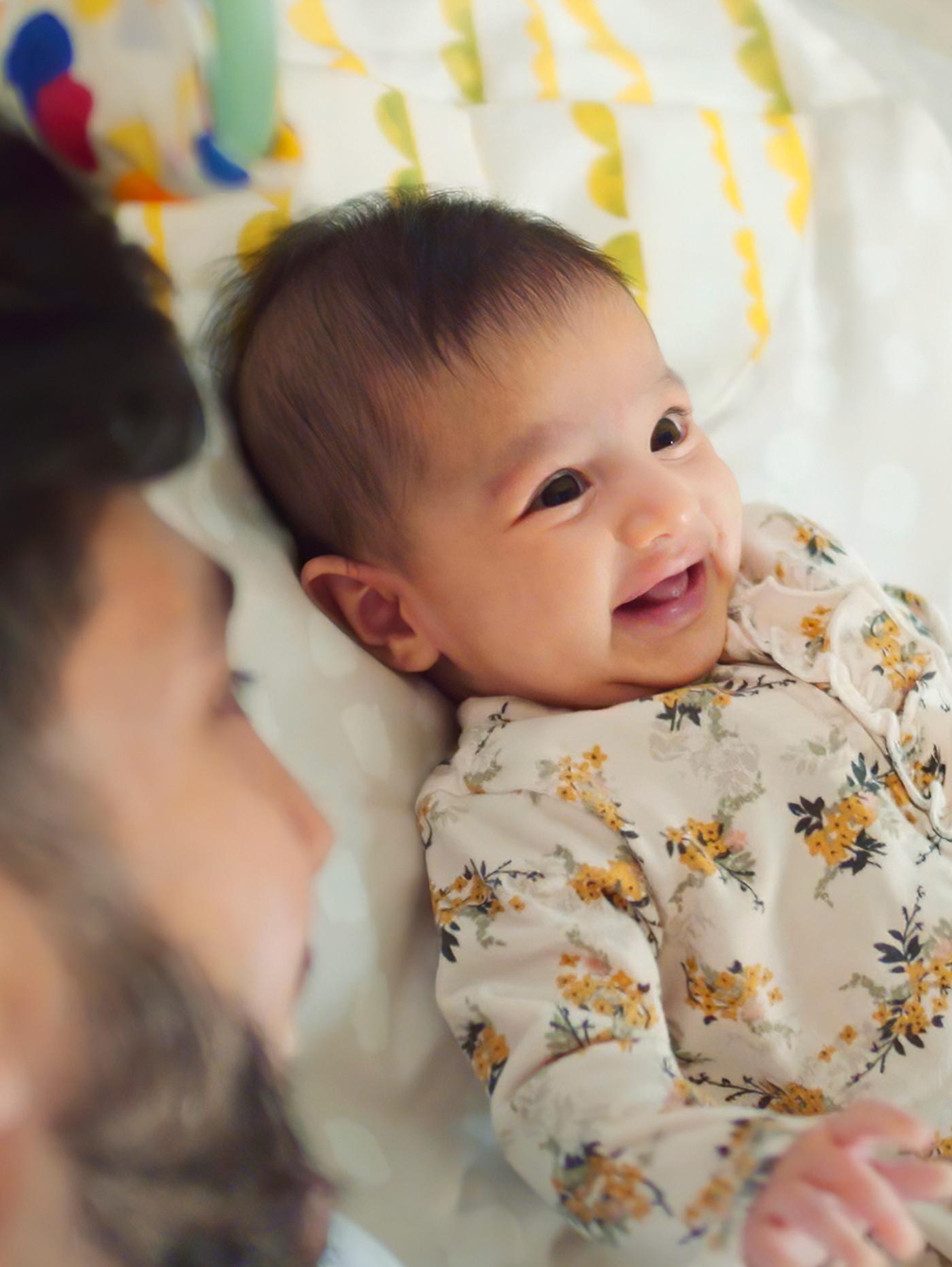 A baby with dark hair and wearing a floral jumpsuit lies on his back and smiles while his Dad looks on.