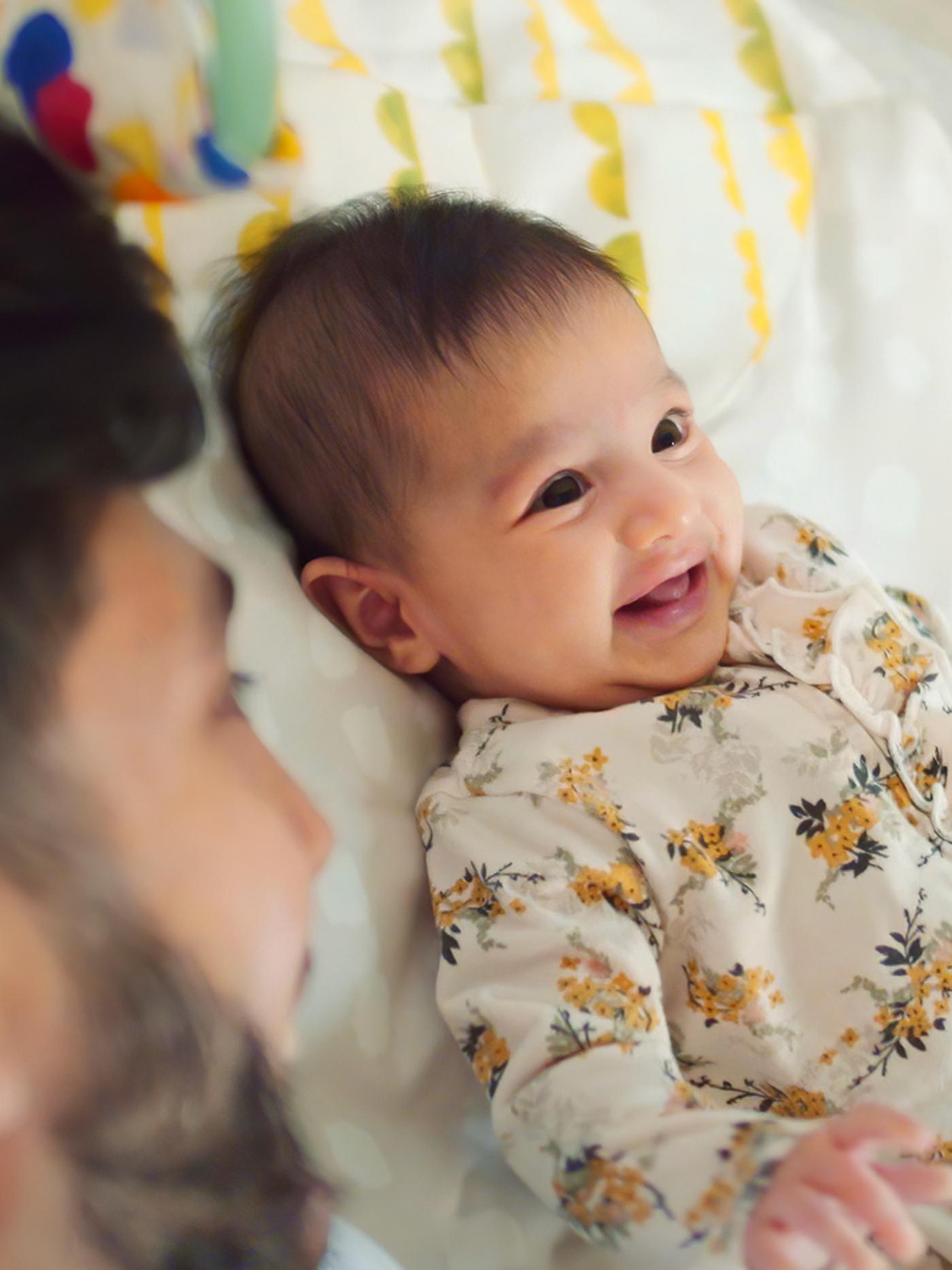 A baby with dark hair and wearing a floral jumpsuit lies on his back and smiles while his Dad looks on.