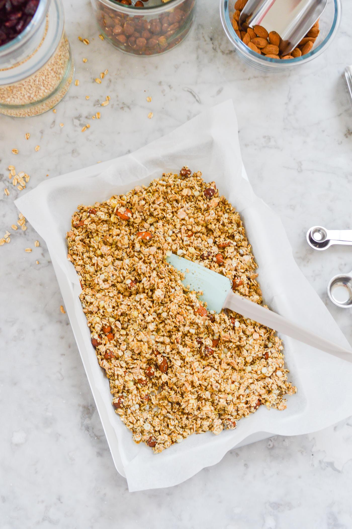A baking tray with unbaked muesli on baking paper, placed on a marble worktop, surrounded by jars of ingredients and a light blue spatula.
