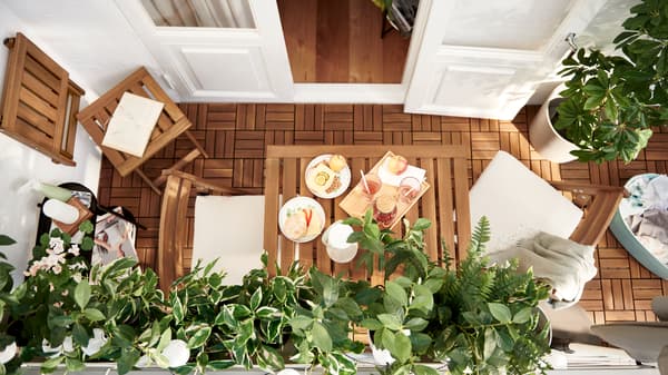 A balcony characterised by slatted acacia – NÄMMARÖ furniture and RUNNEN floor decking – and leafy FEJKA artificial plants.
