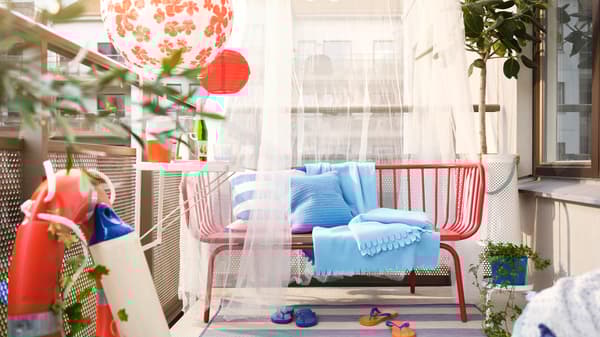 A balcony with a red BRUSEN outdoor sofa with blue cushions and throws, a TORPARÖ balcony table and solar powered LED lamps.