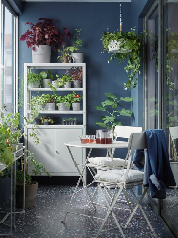 A balcony with a white table and two chairs, some white storage holding plants and plant pots, and a hanging plant.