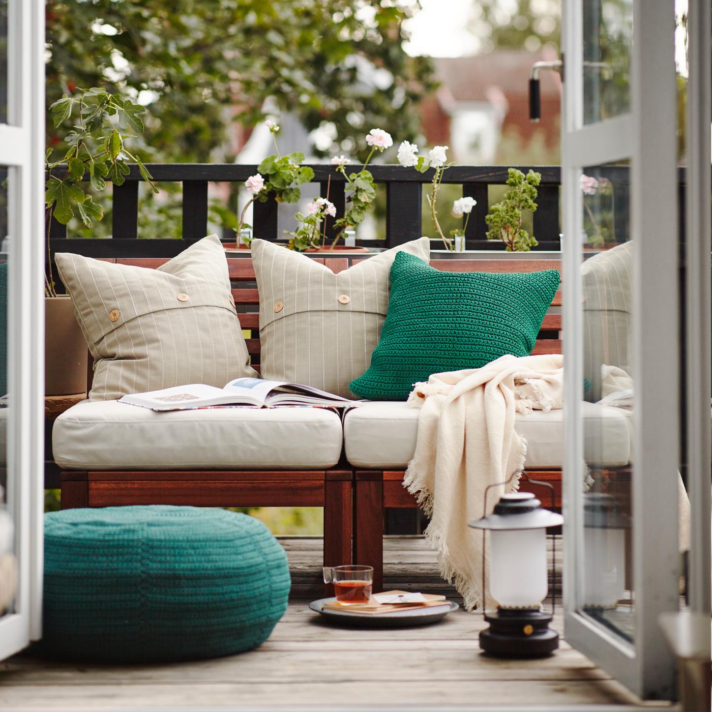 A balcony with a wooden outdoor sofa with white and green cushions, a throw and an open book on it, green plants behind.