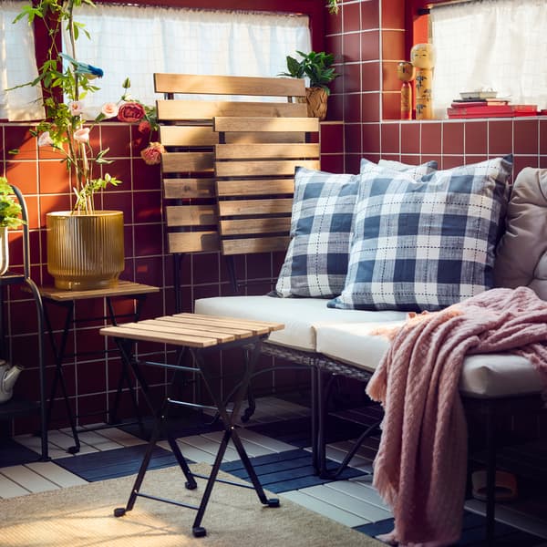 A balcony with dark grey JUTHOLMEN easy chair and light grey-beige cushions surrounded by patterned pillows and green plants.