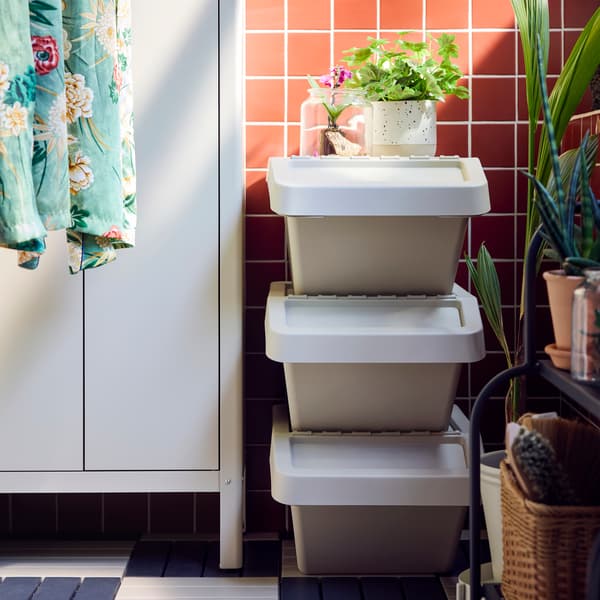 A balcony with three beige SORTERA sorting bins with lid stacked on top of each other against a pink tiled wall.