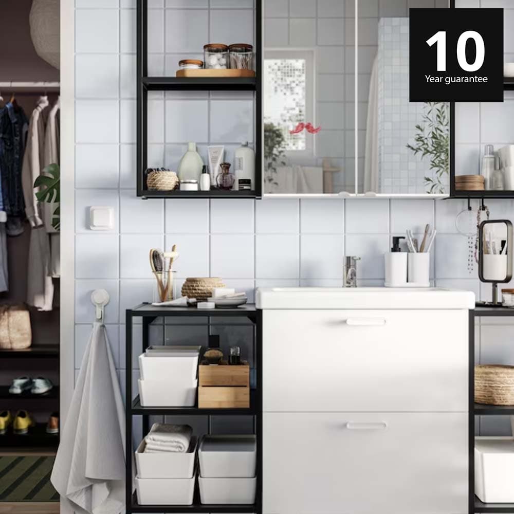 A bathroom has a white ENHET base cabinet with legs, black base frames with shelves and legs and a mirror glass door cabinet. 10-year guarantee logo on top right corner.