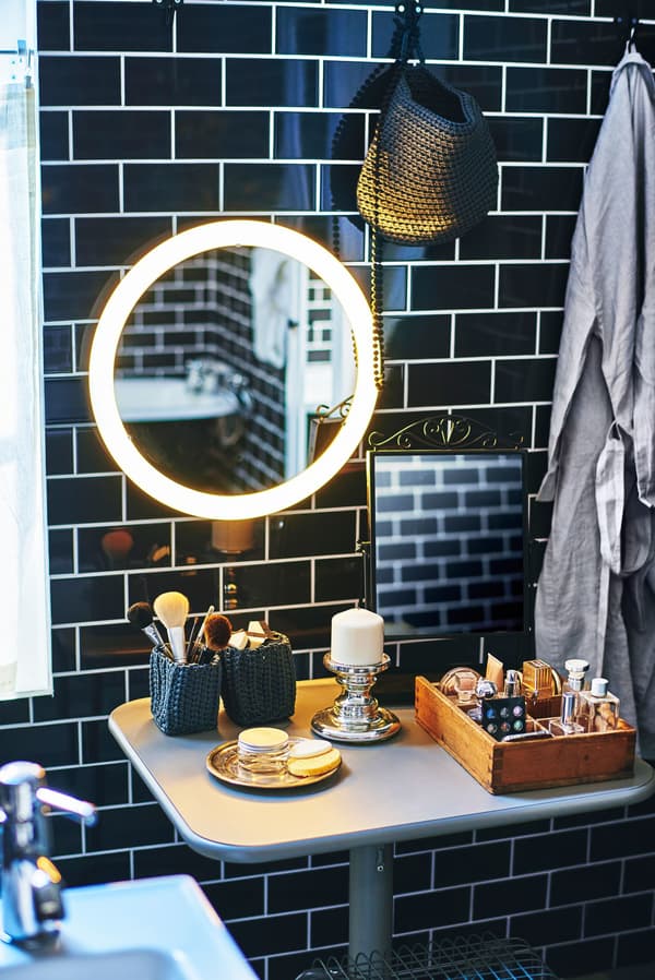A bathroom make-up area under a lighted mirror in a black tiled bathroom.