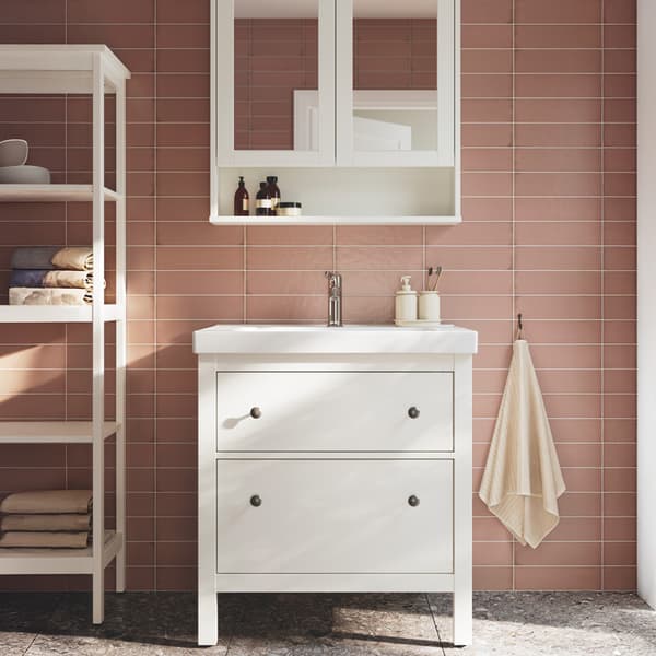 A bathroom setting with pink subway wall tiles with a white HEMNES drawer with a white ORRSJÖN wash-basin with tap leaned against the wall with a white HEMNES mirror cabinet with 2 door and a open shelving for essentials. 