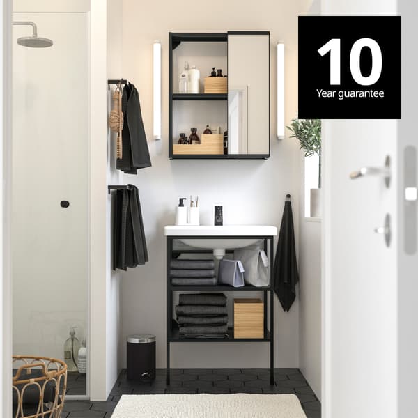 A bathroom setting with white walls and dark grey hexagon toilet tiles with a ENHET base frame with legs and a white wash basin and a mirror glass door cabinet mounted on the wall above it. A 10-year guarantee logo on top right corner.