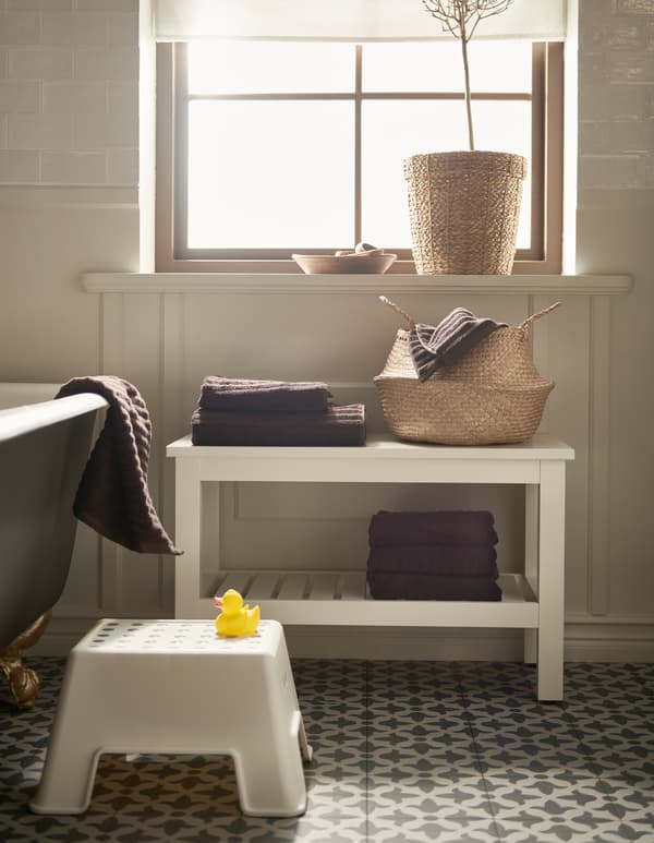 A bathroom with a rattan basket, a white BOLMEN step stool, and a white HEMNES bench with a shelf for towels.