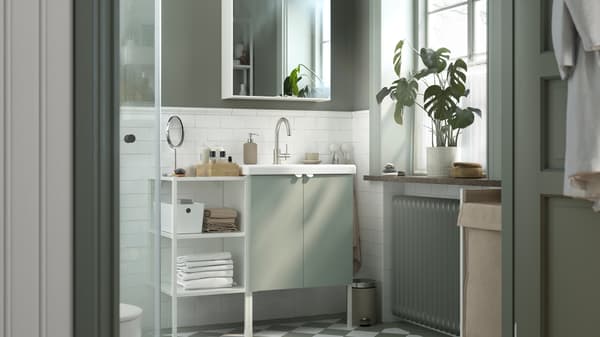 A bathroom with a TVÄLLEN washbasin and ENHET doors in pale grey-green and ENHET wall shelves in white with towels and items.