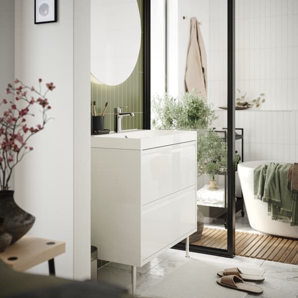 A bathroom with an IKEA ÄNGSJÖN washbasin vanity unit in white with integrated handles, a bathtub behind it and a small bathroom shelf with castors