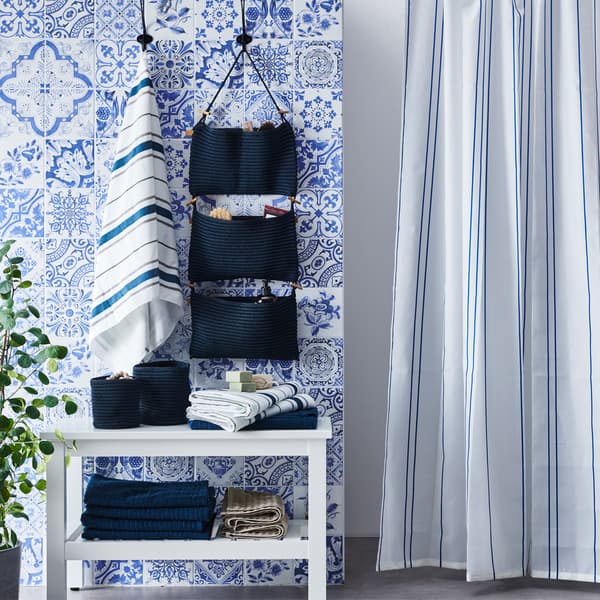 A bathroom with blue and white patterned wall tiles, striped shower curtain, hanging storage and towels folded on a storage unit.