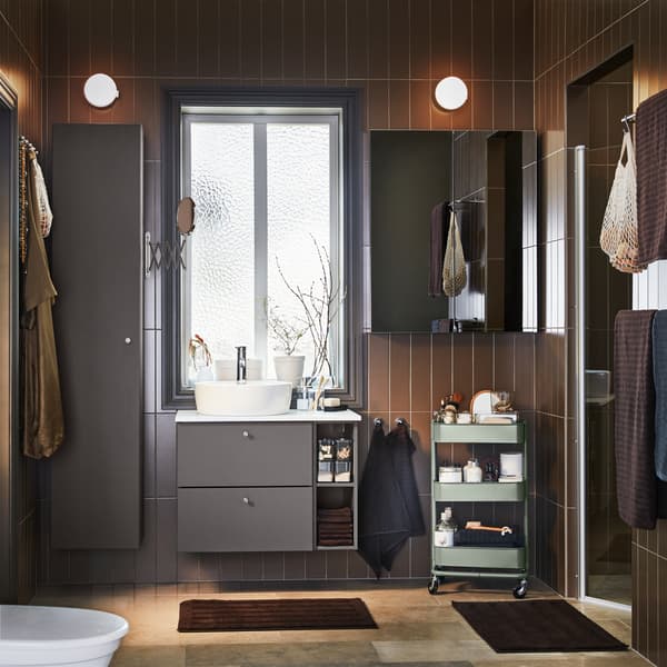 A bathroom with dark grey bathroom furniture, a cabinet with mirror door, a grey-green trolley and dark brown bath mats.