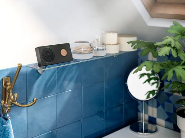 A battery powered IKEA ENEBY black portable Bluetooth speaker on a glass shelf above blue tiles in a bathroom.