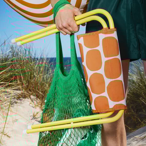 A beach scene with a person holding a bright yellow STRANDÖN folding stool and a bright green bag, ready for a day out.