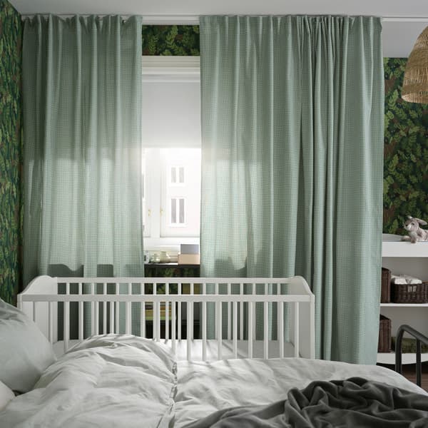 A bed and crib stand by a window with green/white curtains and a white block-out roller blind that covers half the window.