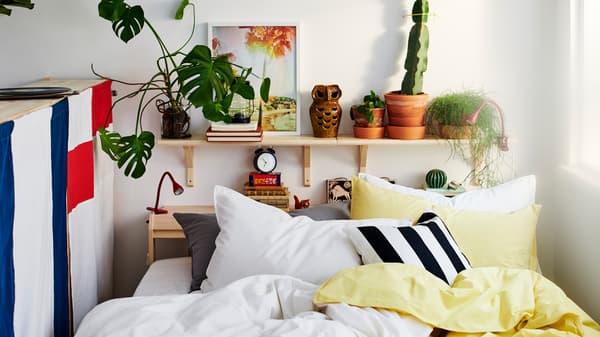 A bed between a window and the back of a storage unit with many pillows and yellow, white, black and grey bed linen.