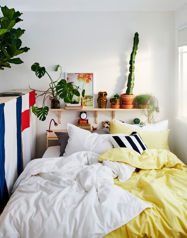 A bed between a window and the back of a storage unit with many pillows and yellow, white, black and grey bed linen.