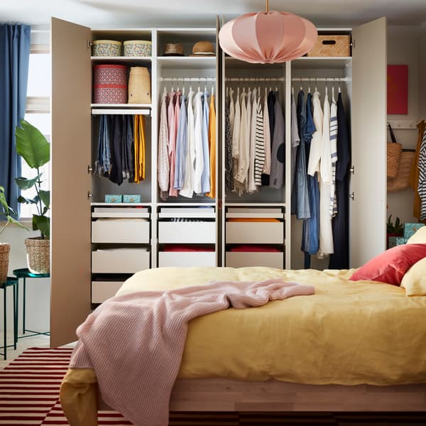 A bed frame in birch, yellow bed textiles, a red/white rug and an open wardrobe with clothes on rods, shelves and in drawers.