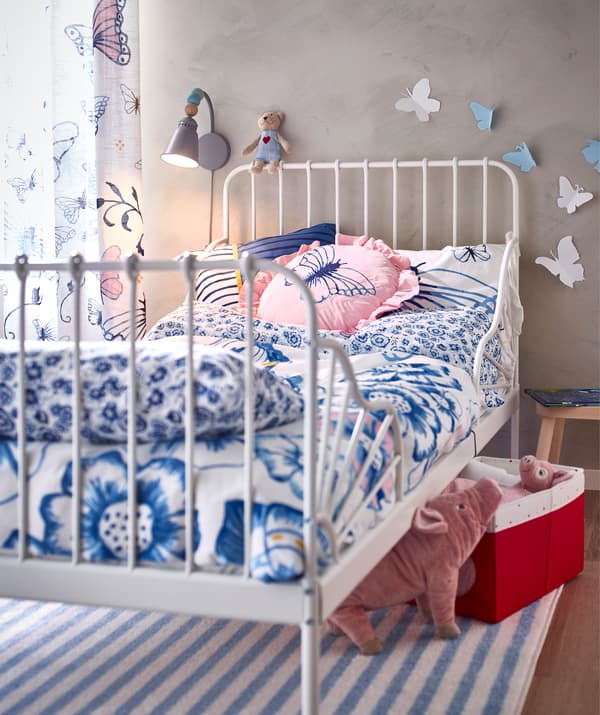 A bed frame with blue and white floral bedding, cushions and curtains with a butterfly motif, and toys under the bed.