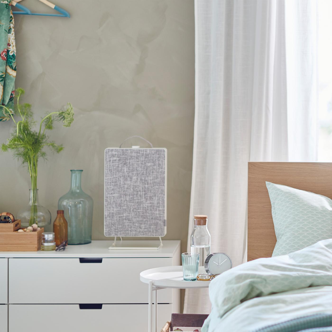 A bed in front of a window and next to a chest of drawers with a FÖRNUFTIG air purifier on top.