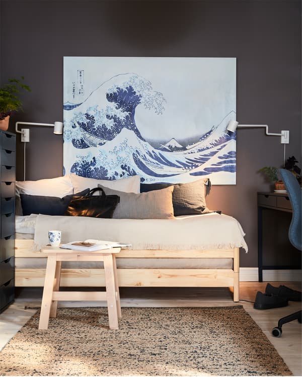 A bed in pine and a birch stool with a book and a mug in front, a big painting with a wave and two white wall-mounted lamps.