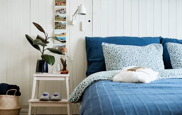 A bed made up with blue bedding, a step stool doubles as a side table, on it is a carafe of infused water and drinking glass.