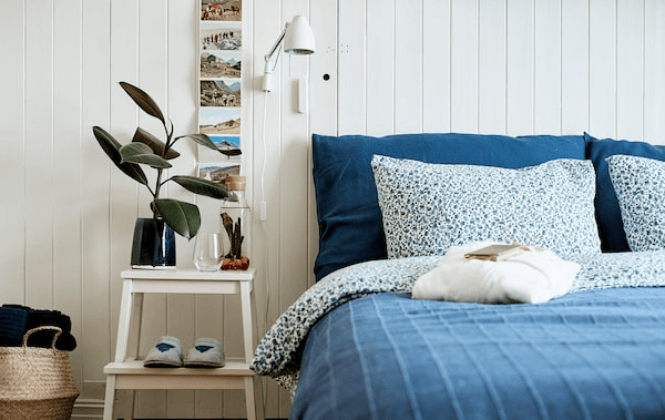 A bed made up with blue bedding, a step stool doubles as a side table, on it is a carafe of infused water and drinking glass.