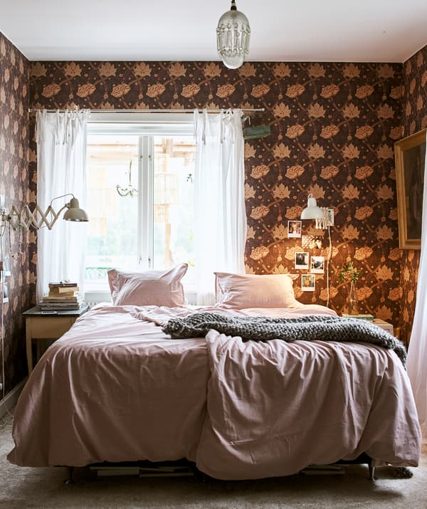 A bed made up with pink bedding and a gray throw in the centre of a room with a chandelier and floral patterned wallpaper.