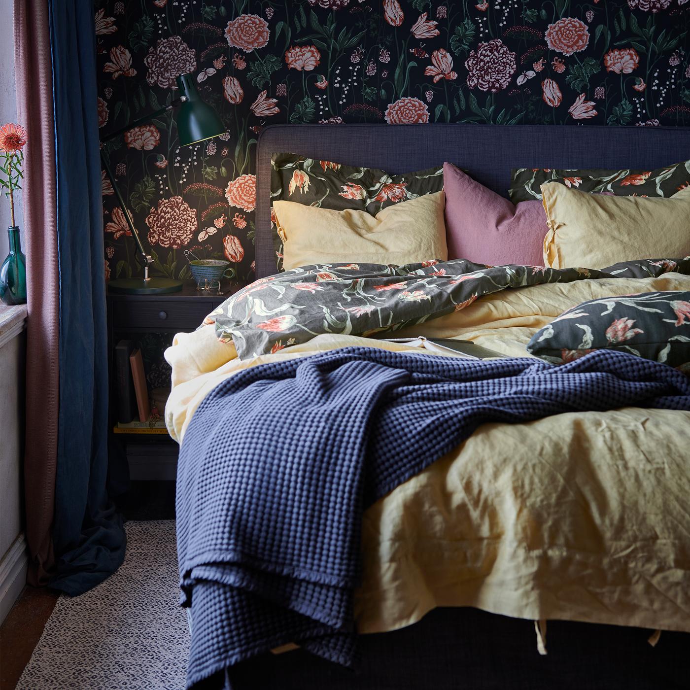 A bed made with yellow, blue and floral bed linen, and a blue folded throw, on a rug by a wall with dark floral pattern.