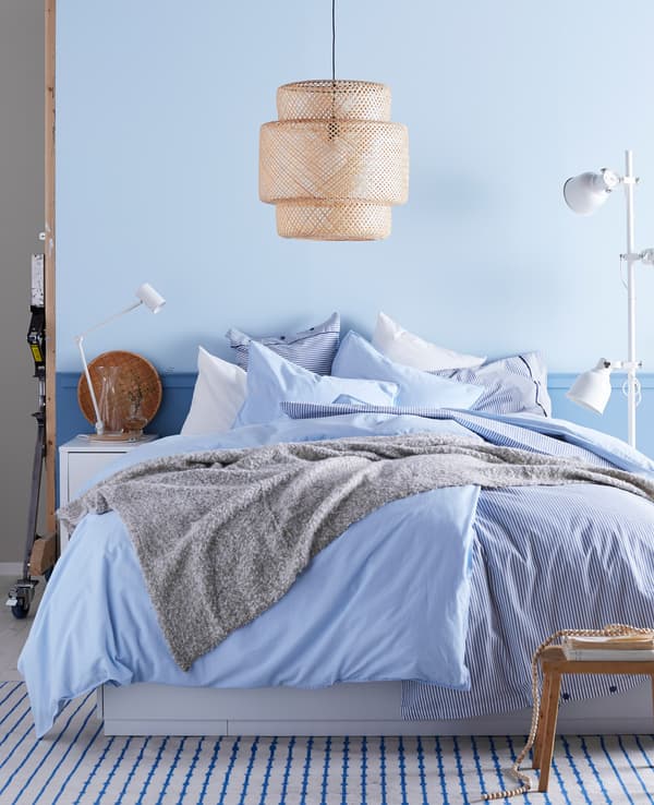 A bed of blue and white linens under a wicker ceiling lamp in a blue bedroom.