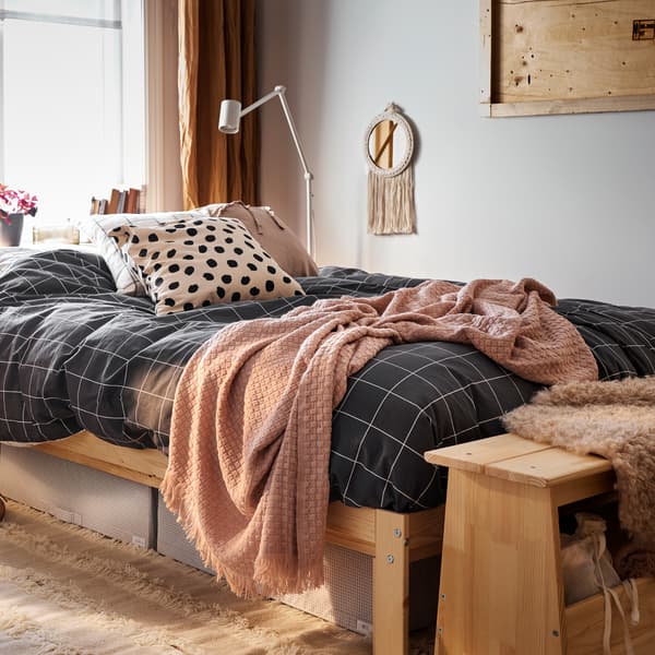 A bed with a pine bed frame made with white black/checked bed linen, plus white/grey storage cases and a rug underneath it.