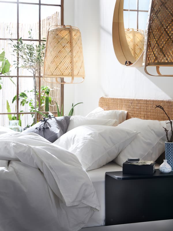 A bed with a rattan headboard, white ÄNGSLILJA quilt cover and pillowcases, pendant lamps in bamboo hang on each side of the bed.