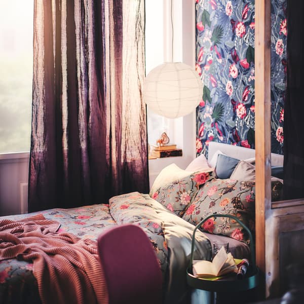 A bed with a throw and bed linen dressed in a NÄSSELKLOCKA duvet cover and pillowcase set, with a pendant lampshade above.