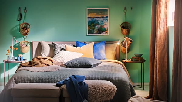 A bed with a white/beige KLEPPSTAD bed frame made with a grey-green bedspread and various cushions, plus two tray tables.