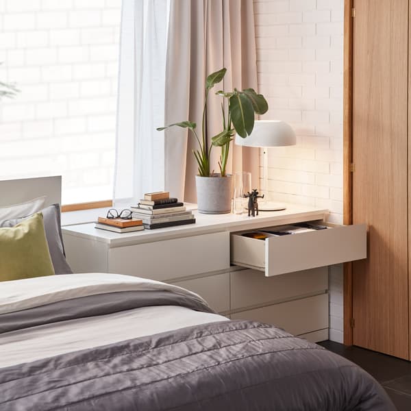 A bed with a white chest of drawers next to it. A green plant, books and a white table lamp stand on the top surface.