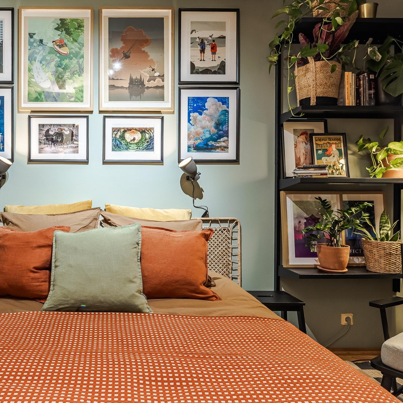 A bed with an orange bedspread and colourful cushions, surrounded by artwork, plants and open shelving.