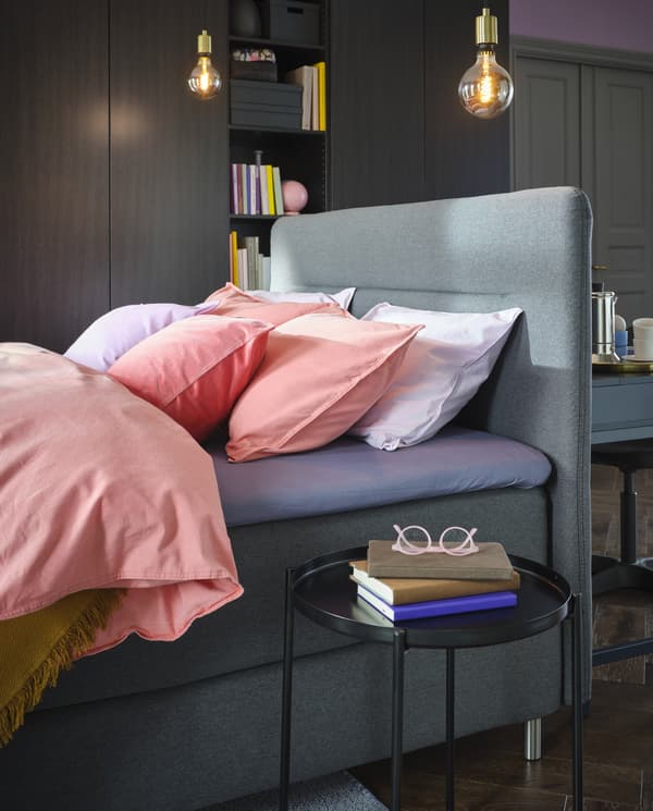 A bed with bed linen in light lilac and light brown-red. Pendant lamps hang above and a black round side table stands beside.