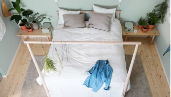 A bed with lightly colored, natural textiles against a light green wall in a symmetrical bedroom with low bedside tables and touches of nature