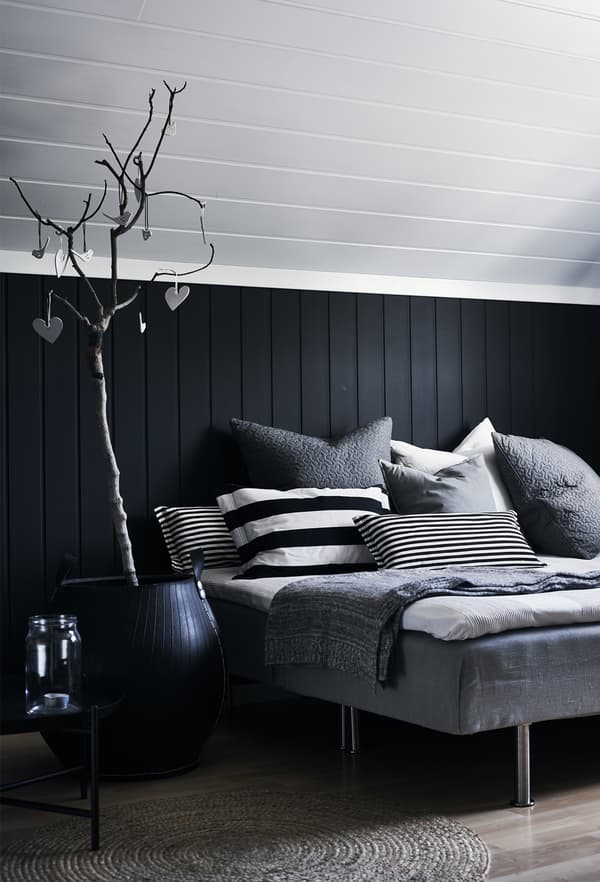 A bed with lots of cushions in different sizes, patterns and grey colours against a black and white shiplap wall.