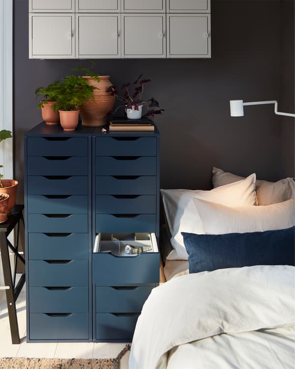 A bed with two tall dark blue drawer units next to it with red-brown plant pots on top. Blue wall-mounted cabinets above.