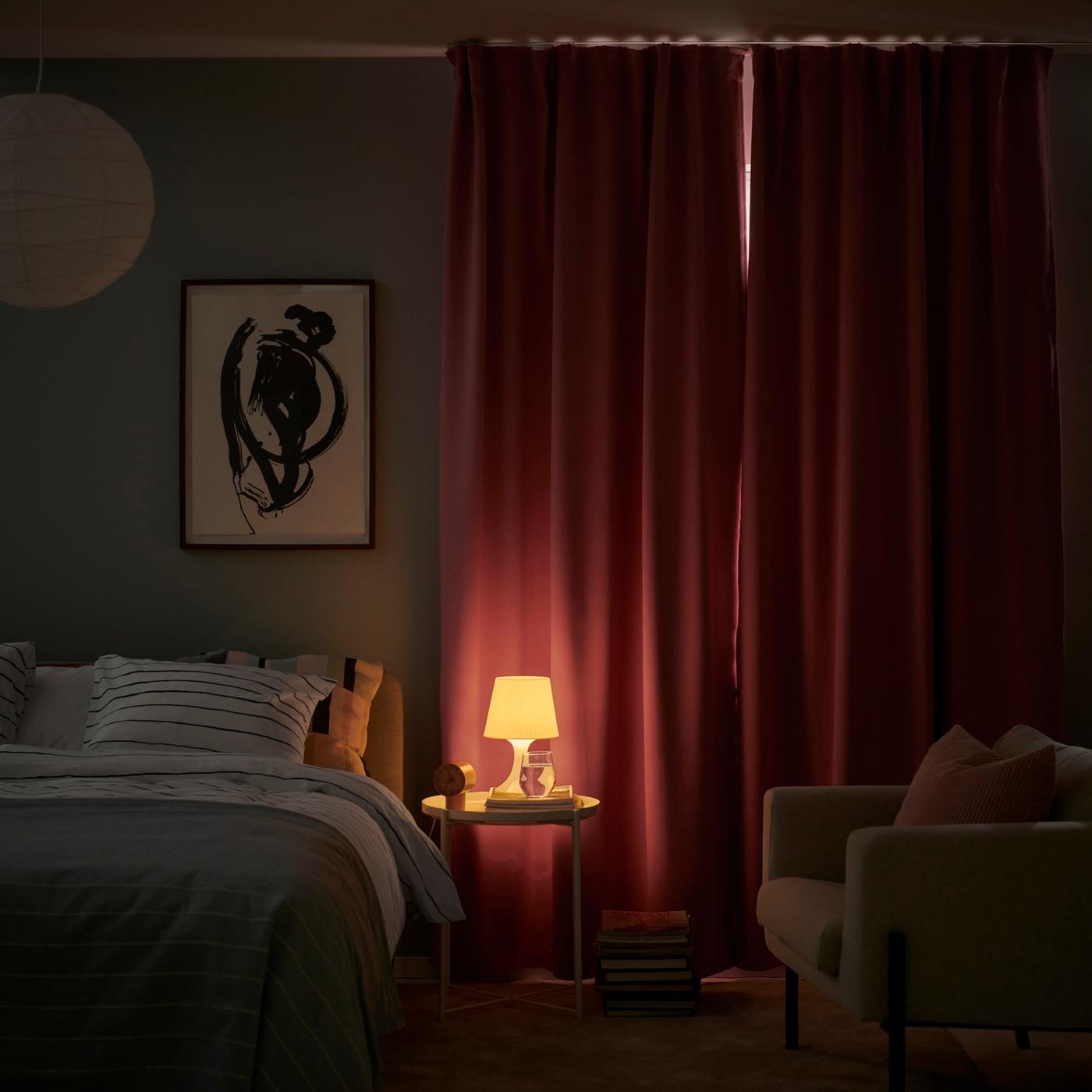 A bedroom at night with deep red curtains drawn across the window and a small bedside table holding a glowing lamp.