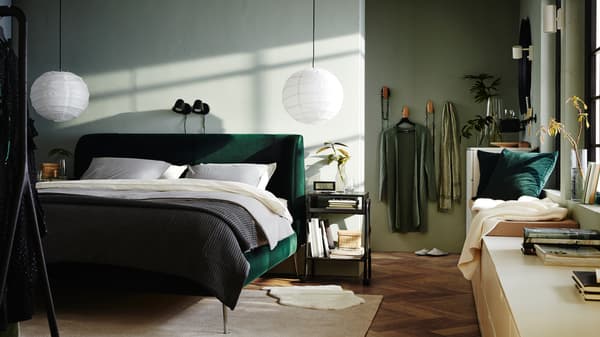 A bedroom containing a green TUFJORD bed with REGOLIT lampshades above and NORDLI drawer units with books by the windows.