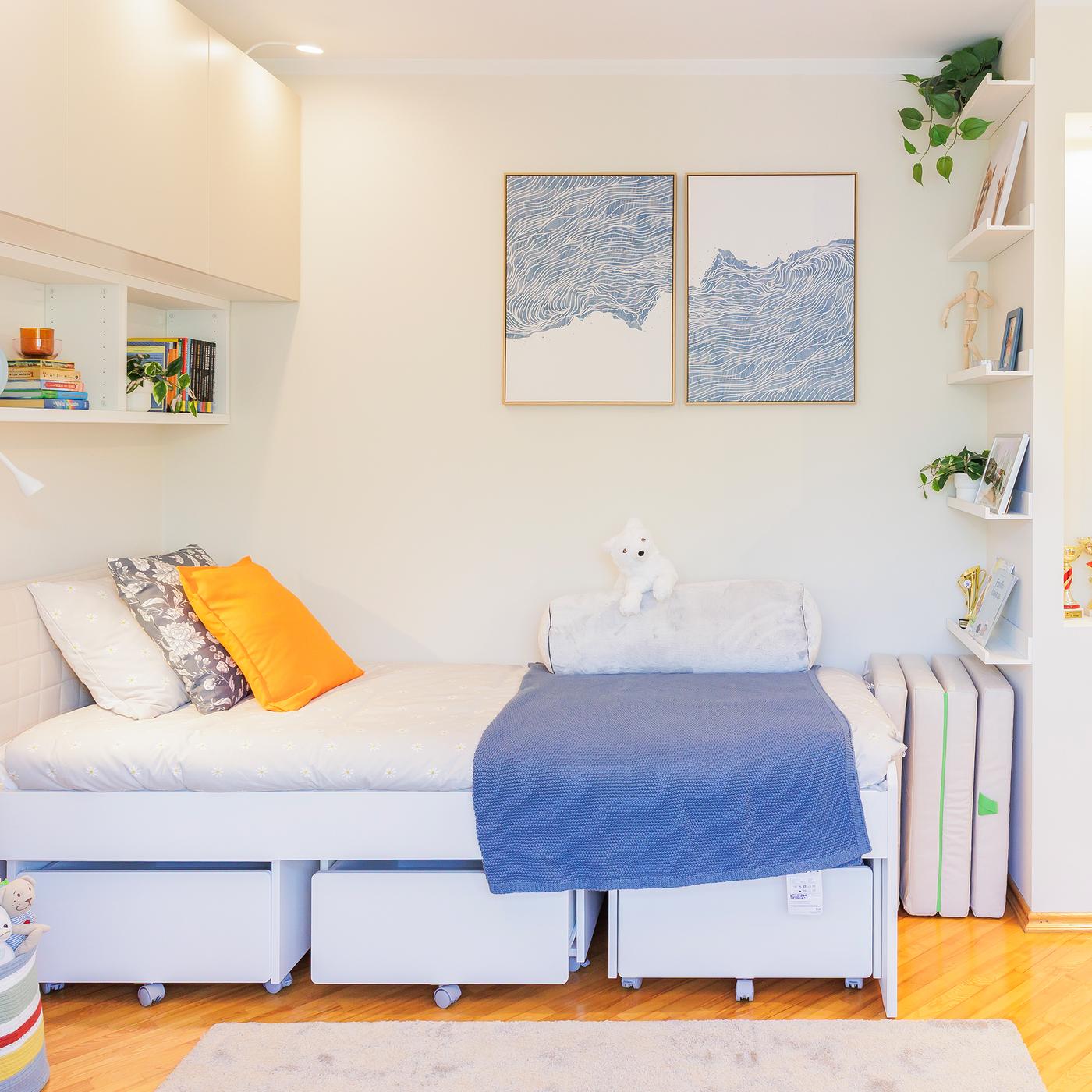 A bedroom corner with a SLÄKT bed frame, white bedding and three storage boxes underneath. Abstract art and some open shelves hang above the bed.