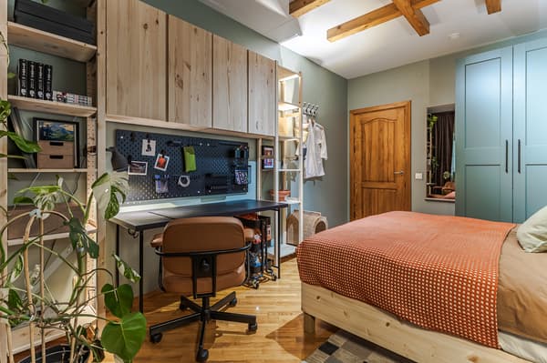 A bedroom featuring a wooden bed with an orange bedspread, a grey-green wardrobe, and a dedicated workspace with a desk, chair and wall storage.